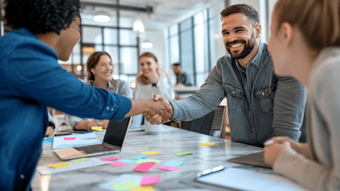 Business professionals shake hands across a table while their teams work together