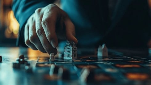 A businessman in a dark blue suit moving a building gamepiece on a game board