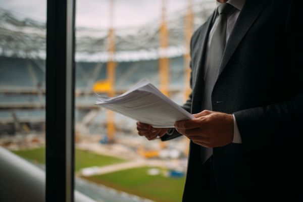 businessman holding papers in front of a window overlooking the construction of a new football stadium