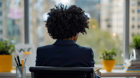 View from behind of a woman wearing a dark blue suit sitting at a desk while writing with a pencil