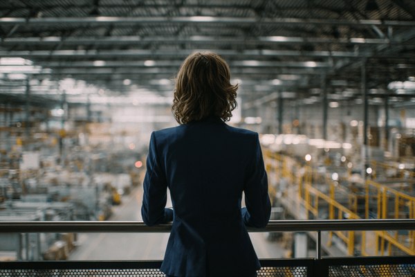 The view from behind a businesswoman in a dark blue suit standing on a catwalk that overlooks a factory floor