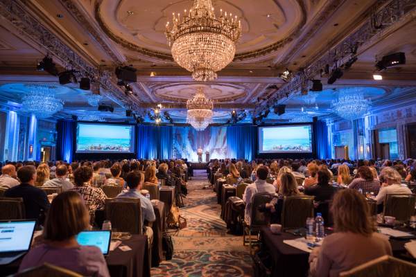 conference attendees sitting in a ballroom watching the keynote speaker on stage next to two large screens