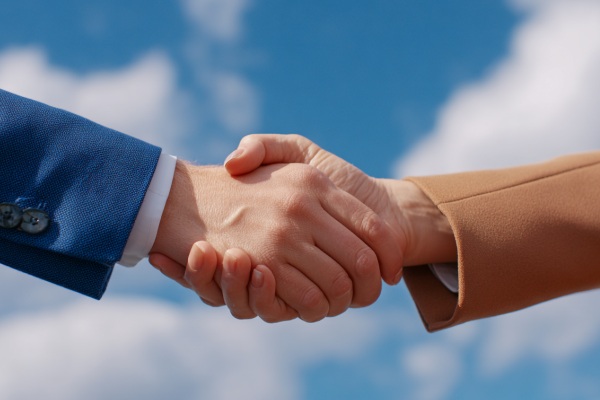 A close-up of a businessman wearing a dark blue suit and a businesswoman wearing a tan suit shaking hands in front of a blue sky with fluffy clouds