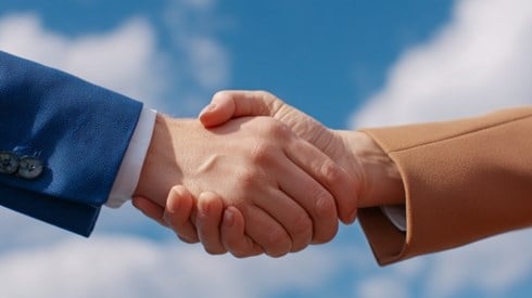 A close-up of a businessman wearing a dark blue suit and a businesswoman wearing a tan suit shaking hands in front of a blue sky with fluffy clouds