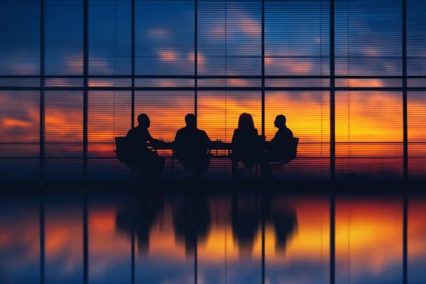 silhouette of four businesspeople sitting at a conference table in front of a glass wall overlooking a sunrise