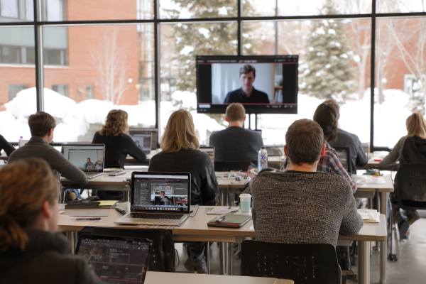 students at desks with laptops watching an instructor on a screen at the front of the room, snow visible outside window