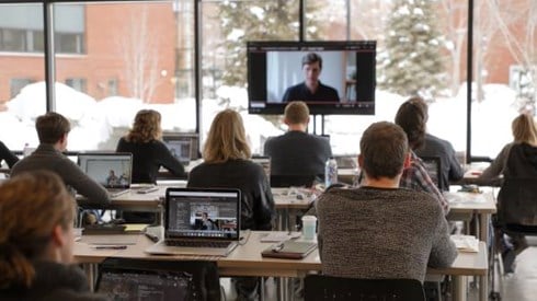 students at desks with laptops watching an instructor on a screen at the front of the room, snow visible outside window