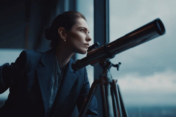 A businesswoman wearing a dark blue suit next to a telescope looking out the window of an office