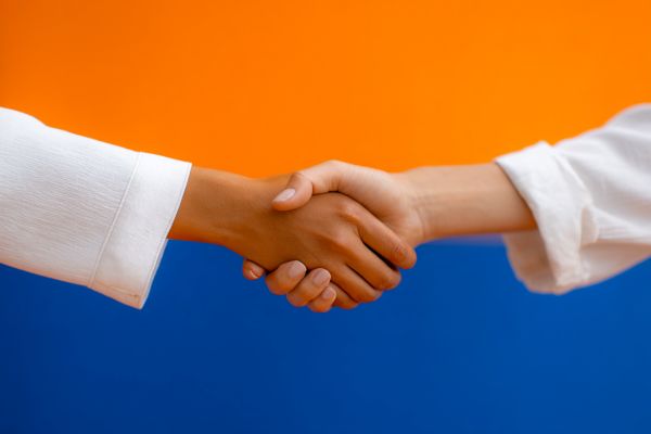 two women's hands with white sleeves shaking over a blue and orange background