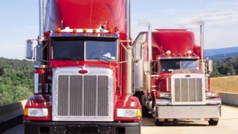 Two red semi-trucks taking up both lanes of the roadway with one in front of the other with trees and hills on each side