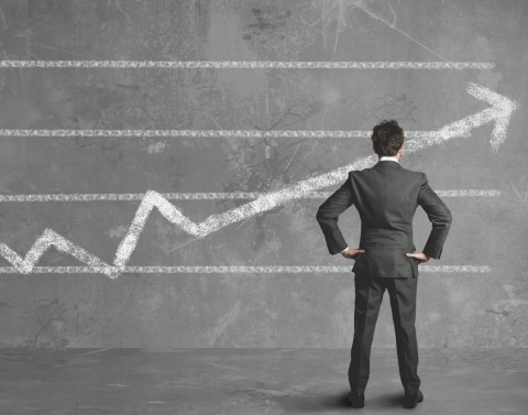 Man in suit standing on gray floor in front of chalkboard wall with graph drawn in chalk and arrow pointing upward