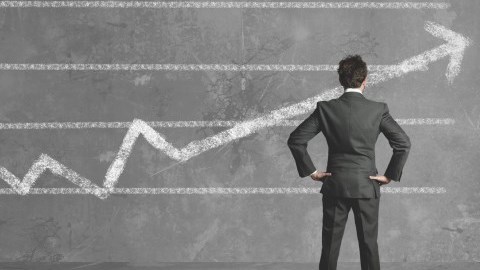 Man in suit standing on gray floor in front of chalkboard wall with graph drawn in chalk and arrow pointing upward