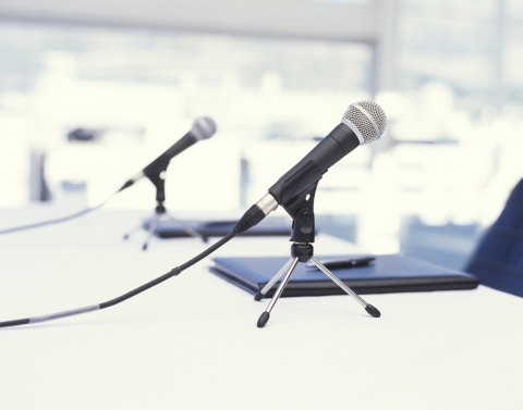 2 tabletop microphones sitting in front of black notebooks with a pen on top at a white table with chair