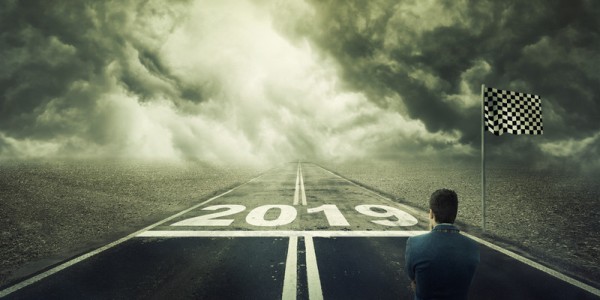 A businessman contemplates a racetrack with a starting line and flag at 2019 and the road ahead is dark with thick clouds