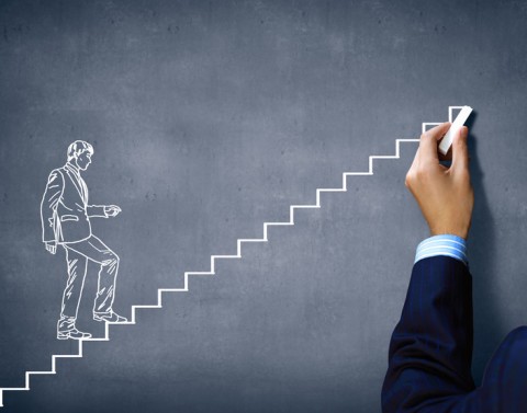 Hand holding chalk and drawing a picture of a man walking up stairs on a chalkboard to indicate promotion