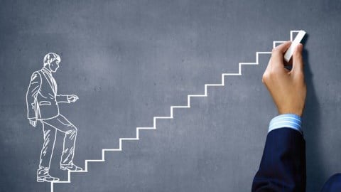 Hand holding chalk and drawing a picture of a man walking up stairs on a chalkboard to indicate promotion