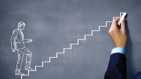 Hand holding chalk and drawing a picture of a man walking up stairs on a chalkboard to indicate promotion