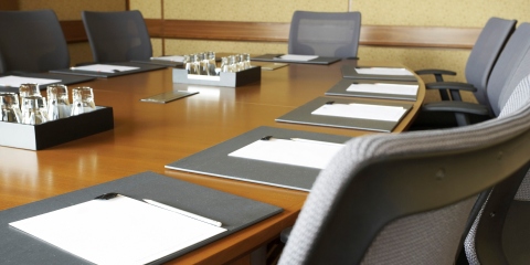 A corporate board room with a long wooden oval table surrounded by gray executive chairs. There are place settings with documents at each chair.