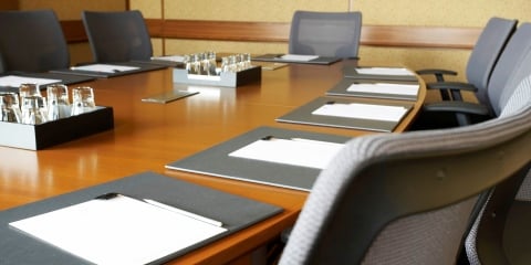 A corporate board room with a long wooden oval table surrounded by gray executive chairs. There are place settings with documents at each chair.