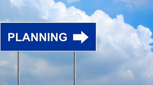 A blue rectangular road sign with the word PLANNING and an arrow pointing to the right in front of sky with white clouds