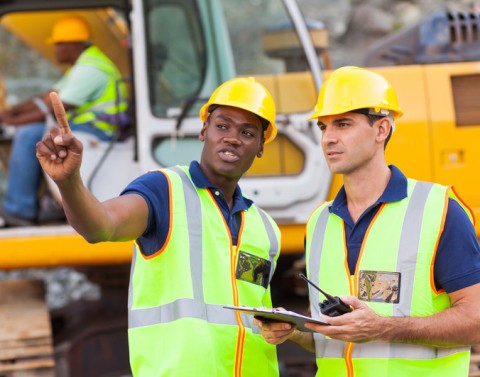 Two construction workers at a site discuss the project and another is operating a large construction vehicle in background