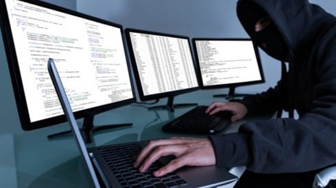 A hooded computer man in front of 3 large monitors with one hand working from a laptop and the other from a computer keyboard