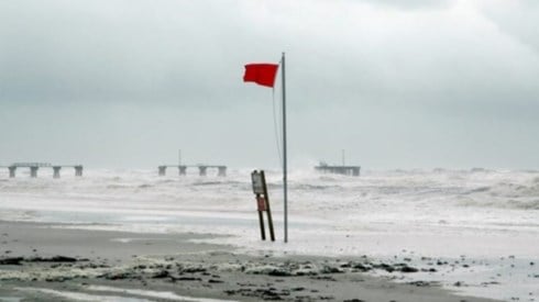 Beach with red warning flag, rough waves, debris, broken pier, and storm clouds