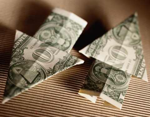 Two one-dollar bills folded in the shape of arrows pointing in opposite directions sitting on brown corrugated cardboard
