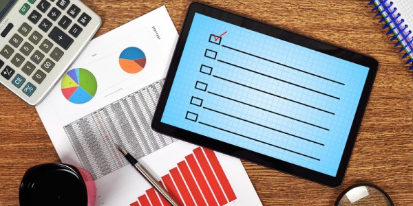 Desk with calculator coffee magnifying glass a page of graphs and a tablet displaying an empty checklist with one checkmark