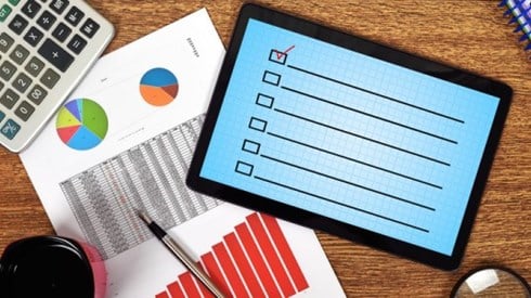 Desk with calculator coffee magnifying glass a page of graphs and a tablet displaying an empty checklist with one checkmark