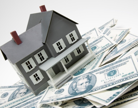 A small scale gray model house with white trim and a front porch sitting on top of a pile of 20-dollar bills