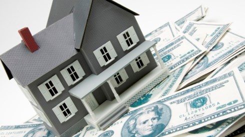 A small scale gray model house with white trim and a front porch sitting on top of a pile of 20-dollar bills
