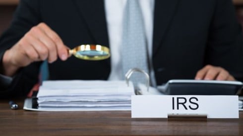 IRS desk sign and man in suit holding a magnifying glass over binder of documents while running a desk calculator