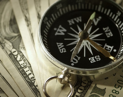 A brass compass having white settings on a black face and brass hands sitting on top of one hundred dollar bills that are fanned out