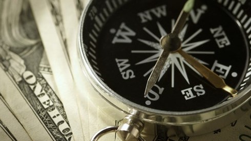 A brass compass having white settings on a black face and brass hands sitting on top of one hundred dollar bills that are fanned out