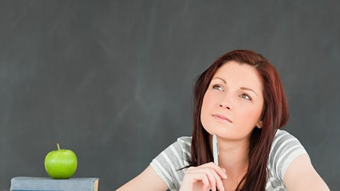 A student thinking and sitting at a desk that has an apple on top of a stack of books alongside other groups of papers