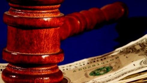A shiny all wood mahogany colored gavel resting on top of a pile of one dollar bills with a blue background