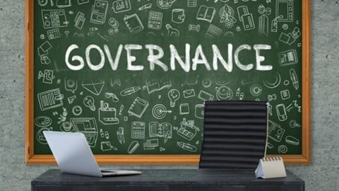 A desk with a calendar and laptop in front of a chalkboard with the word GOVERNANCE and different learning tools drawn on it