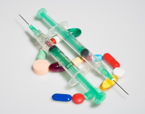 Two syringes with needles resting on top of pills forming the shape of a dollar sign