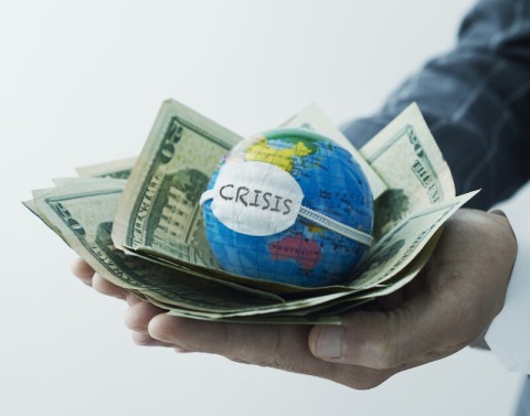 Hand holding globe with face mask on it on top of pile of dollar bills