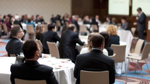Business professionals seated at round tables in a conference hall listen to a speaker give a presentation.