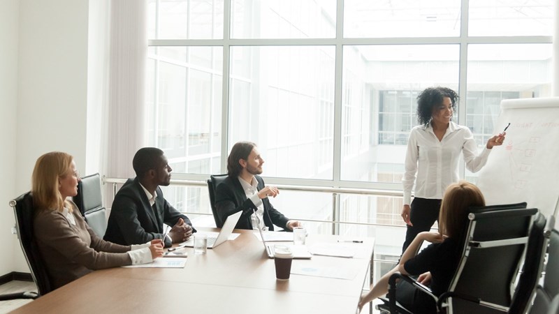 Directors Strategize with Laptops around a Conference Table with Discussion Leader Illustrating on Flipchart