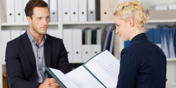 Woman holding a resume and interviewing a man sitting in an office with binders on shelves in the background