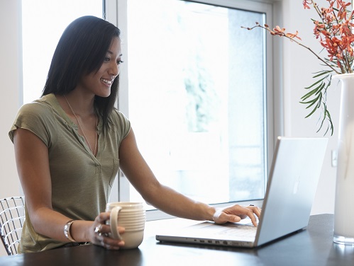 A young woman sitting at desk holding a mug with a laptop open and a vase with flowers in it and a window behind her.