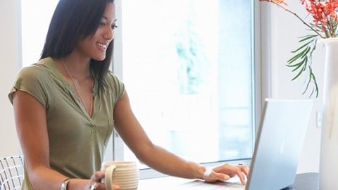 A young woman sitting at desk holding a mug with a laptop open and a vase with flowers in it and a window behind her.