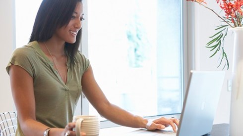 A young woman sitting at desk holding a mug with a laptop open and a vase with flowers in it and a window behind her.