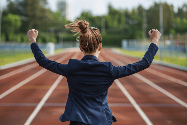 Businesswoman in a dark blue suit winning a race on a track and holding her hands up in victory