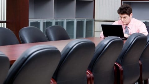 Man sitting at the end of a conference table in a large room working on a laptop