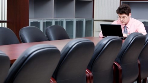 Man sitting at the end of a conference table in a large room working on a laptop
