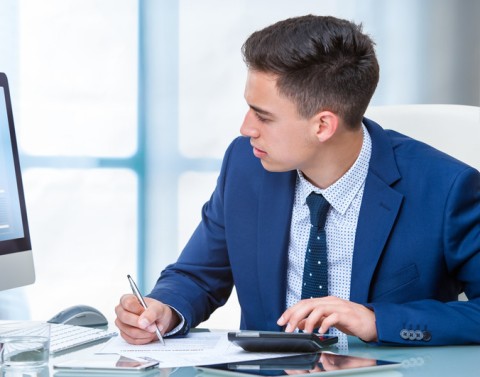 Businessman with paper, pen, and calculator in front of a computer monitor